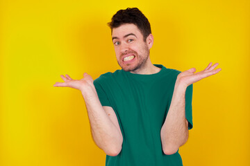Clueless young Caucasian man wearing green T-shirt against yellow wall shrugs shoulders with...
