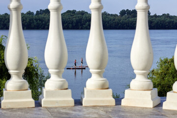 Two women rowing on stand up paddle boarding (SUP) along the calm summer Danube river. View through the columns