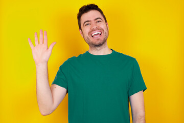 young Caucasian man wearing green T-shirt against yellow wall Waiving saying hello happy and smiling, friendly welcome gesture. © Jihan