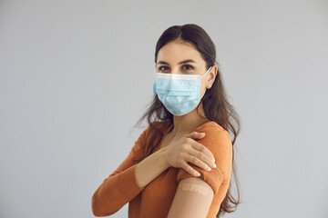 Happy beautiful young woman wearing medical face mask isolated on gray copy space background shows adhesive plaster on arm after getting safe Covid 19 vaccine. Coronavirus vaccination campaign concept
