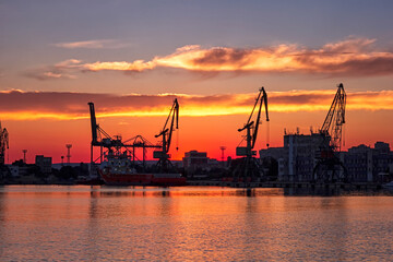 Fototapeta premium Silhouettes of port cranes at stunning red sunset. Cargo ship terminal at the twilight scene.