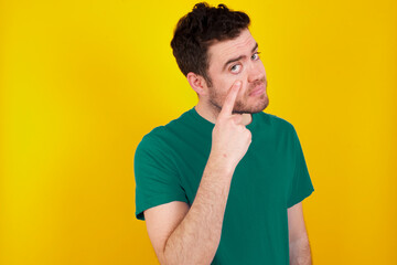 young Caucasian man wearing green T-shirt against yellow wall Pointing to the eye watching you gesture, suspicious expression.