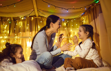 Happy family spend fun time together at home lifestyle. Overjoyed mother playing with adorable children in tent with glowing garlands light decoration at evening. Cheerful kids and loving mom