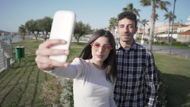 Young caucasian couple of tourists on vacation at sea ocean resort use smartphone to chat live with distant friends. Two bloggers film themselves with smartphone sharing journey on social vlog network