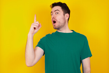 young Caucasian man wearing green T-shirt against yellow wall pointing finger up and looking inspired by genius thought, showing good idea sign, having clever solution in mind