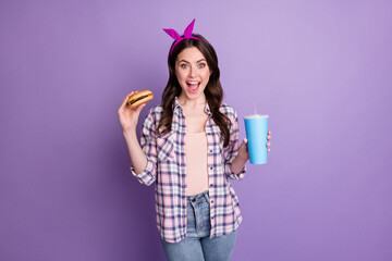 Portrait of attractive cheerful girl drinking soda eating tasty yummy beef burger isolated over bright violet purple color background