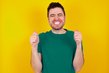 young Caucasian man wearing green T-shirt against yellow wall being excited for success with raised arms and closed eyes celebrating victory. Winner concept.