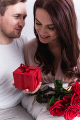 Smiling woman holding gift box and roses near husband in bedroom