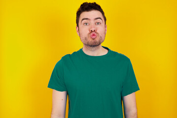 Shot of pleasant looking young handsome caucasian man wearing green t-shirt against yellow background , pouts lips, looks at camera, Human facial expressions