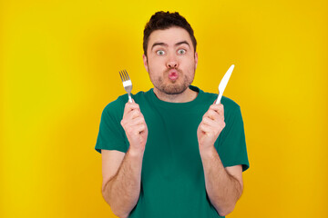 Model hungry young handsome caucasian man wearing green t-shirt against yellow background holding in hand fork knife want tasty yummy pizza pie