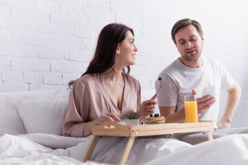 Woman looking at husband with smartphone near breakfast on blurred foreground on bed