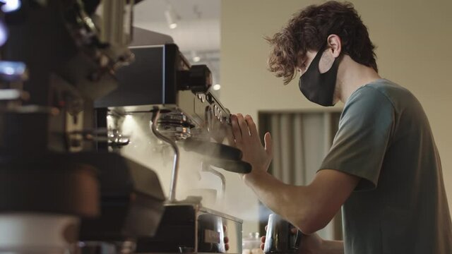 Lockdown of young male barista wearing t-shirt and mask preparing milk for cappuccino using modern automatic coffee machine in cafe