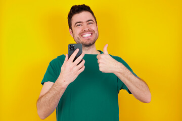 Portrait of young handsome caucasian man wearing green t-shirt against yellow background using and texting with smartphone  happy with big smile doing ok sign, thumb up with fingers, excellent sign