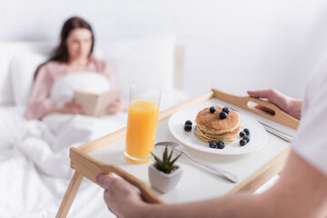Obraz na płótnie Canvas Man holding breakfast on tray near wife on blurred background in bedroom