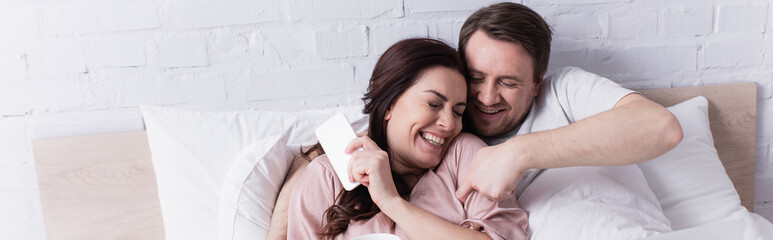 Smiling man tickling wife with smartphone on bed, banner - obrazy, fototapety, plakaty