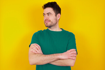 Pleased young handsome caucasian man wearing green t-shirt against yellow background keeps hands crossed over chest looks happily aside