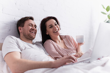 Smiling man using laptop on blurred foreground near wife with cup on bed