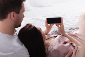 Smartphone with blank screen in hands of woman lying near husband on bed