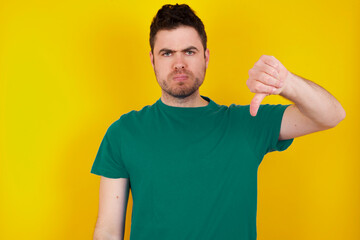 young handsome caucasian man wearing green t-shirt against yellow background looking unhappy and angry showing rejection and negative with thumbs down gesture. Bad expression.