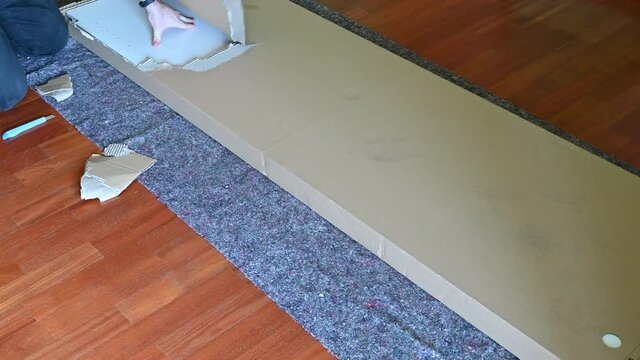 Housework: assembling a piece of furniture. The package has been lying on the ground on a protective sheet, the caucasian man's hands are opening the package by tearing the cardboard.