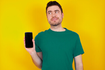 young handsome caucasian man wearing green t-shirt against yellow background holds new mobile phone and looks mysterious aside shows blank display of modern cellular