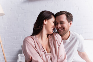 Positive woman in pajama sitting near husband with closed eyes on bed