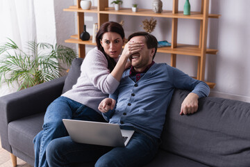 Scared woman covering eyes to husband pointing at laptop on couch