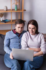 Fototapeta premium Man looking at laptop near cheerful wife on couch in living room