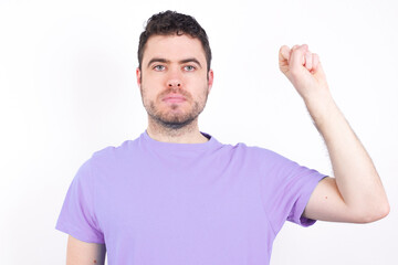 Naklejka premium young handsome caucasian man wearing purple t-shirt against white background feeling serious, strong and rebellious, raising fist up, protesting or fighting for revolution.