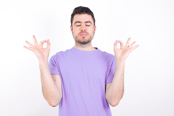 Fototapeta premium young handsome caucasian man wearing purple t-shirt against white background doing yoga, keeping eyes closed, holding fingers in mudra gesture. Meditation, religion and spiritual practices.