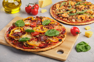 Two homemade pizzas with mozzarella cheese, tomatoes, mushrooms, salami and seafood decorated with basil and arugula on a gray background. Close-up, horizontal orientation without people.