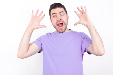 Fototapeta premium young handsome caucasian man wearing purple t-shirt against white background looks with excitement at camera, keeps hands raised over head, notices something unexpected, reacts on sudden news.