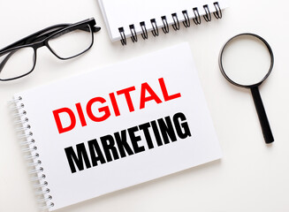 DIGITAL MARKETING is written in a white notebook on a light background near the notebook, black-framed glasses and a magnifying glass.