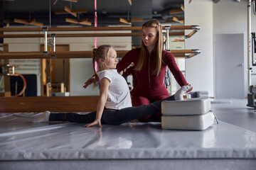 Young confident female teacher is helping girl with stretching in modern hall