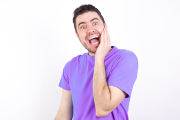 Shocked, astonished young handsome caucasian man wearing purple t-shirt looking surprised in full disbelief wide open mouth with hand near face. Positive emotion facial expression body language.