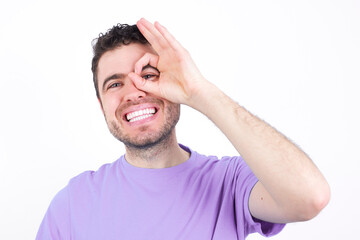 young handsome caucasian man wearing purple t-shirt against white background with happy face smiling doing ok sign with hand on eye looking through finger.