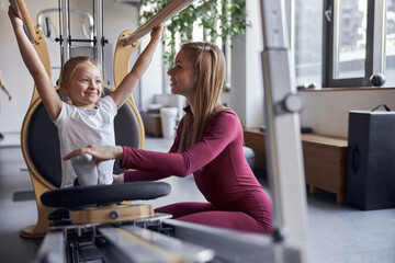 Young confident female teacher is helping girl with stretching in modern hall