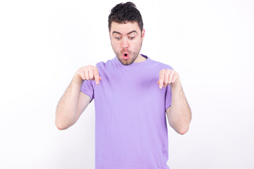 Amazed young handsome caucasian man wearing purple t-shirt against white background points down...