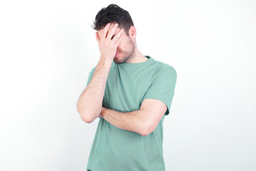 young handsome caucasian man wearing green t-shirt against white background making facepalm gesture while smiling amazed with stupid situation.