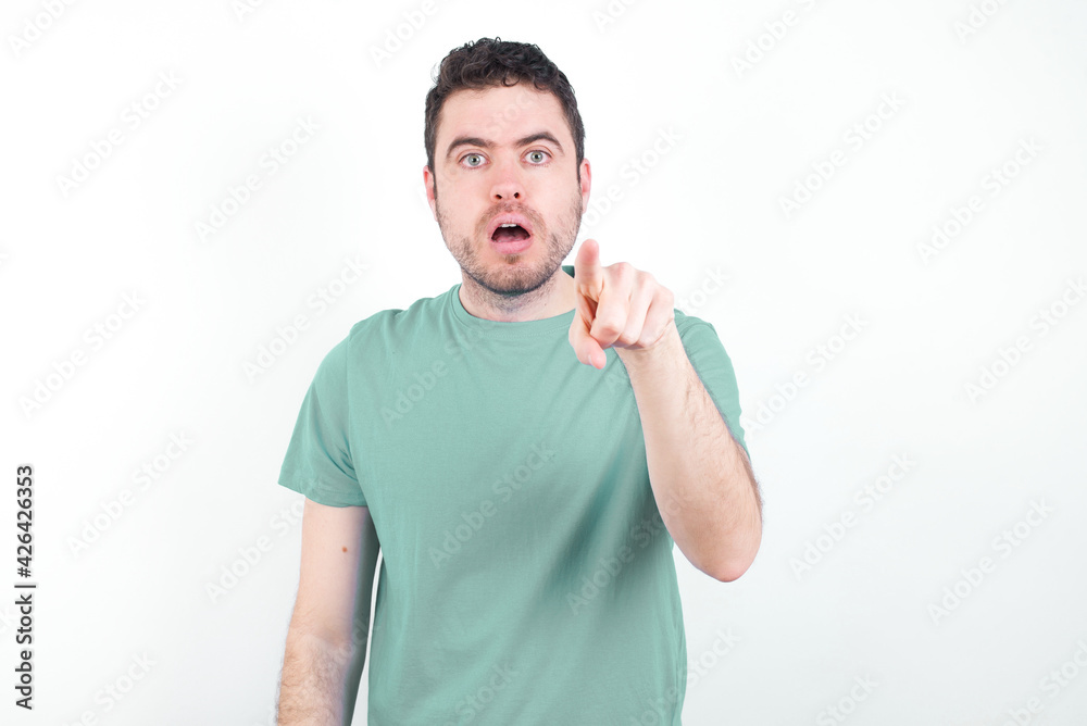 Poster Shocked young handsome caucasian man wearing green t-shirt against white background points front with index finger at camera and. Surprise and advertisement concept.