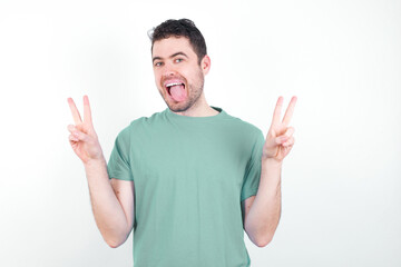 Fototapeta premium young handsome caucasian man wearing green t-shirt against white background with optimistic smile, showing peace or victory gesture with both hands, looking friendly. V sign.