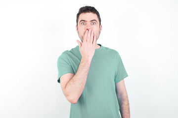 Oh! I think I said it! Close up portrait young handsome caucasian man wearing green t-shirt against white background cover open mouth by hand palm, look at camera with big eyes.
