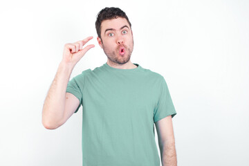 Obraz premium Shocked young handsome caucasian man wearing green t-shirt against white background shows something little with hands, demonstrates size, opens mouth from surprise. Measurement concept.