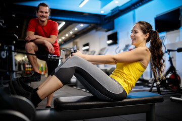 Fototapeta na wymiar Smiling and happy young Caucasian woman exercising on a rowing machine in indoor gym with her boyfriend standing and cheering her in the background