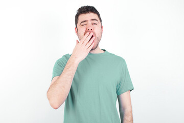 Sleepy young handsome caucasian man wearing green t-shirt against white background yawning with messy hair, feeling tired after sleepless night, yawning, covering mouth with palm.