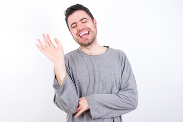 Obraz premium Overjoyed successful young handsome caucasian bearded man wearing pyjama over white wall raises palm and closes eyes in joy being entertained by friends