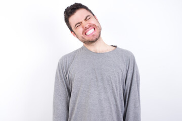 Fototapeta premium Positive young handsome caucasian bearded man wearing pyjama over white wall with overjoyed expression closes eyes and laughs shows white perfect teeth