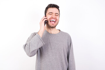 Funny young handsome caucasian bearded man wearing pyjama over white wall laughs happily, has phone conversation, being amused by friend, closes eyes.