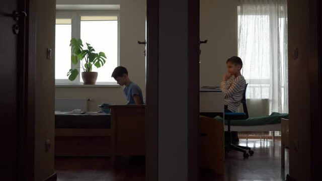 Teenage boy sitting and doing homework with laptop computer. Brother using digital tablet in other bedroom. View through open doors to two rooms from hallway. Online education, entertainment at home