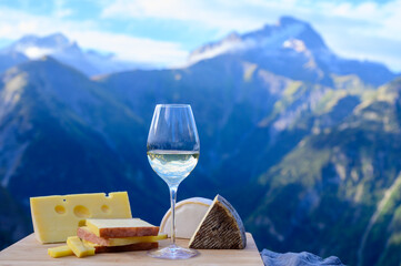 Tasty cheese and wine from Savoy region in France, beaufort, abondance, emmental, tomme and reblochon de savoie cheeses and glass of white wine served outdoor with Alpine mountains peaks on background